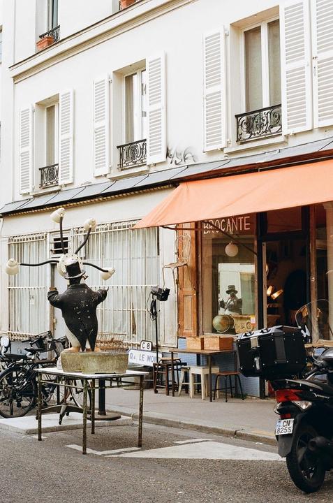 A street in front of a building with an orange awning features a statue of a man in a top hat and a scooter.