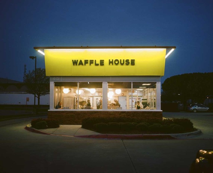 A Waffle House restaurant is illuminated at night, with people visible inside through its large windows.