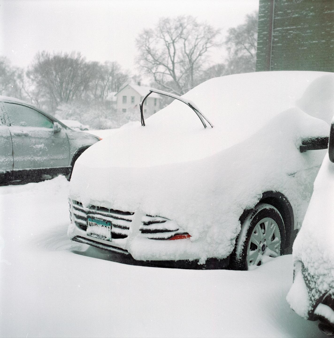 A car is parked on a snowy street with its windshield wipers raised.
