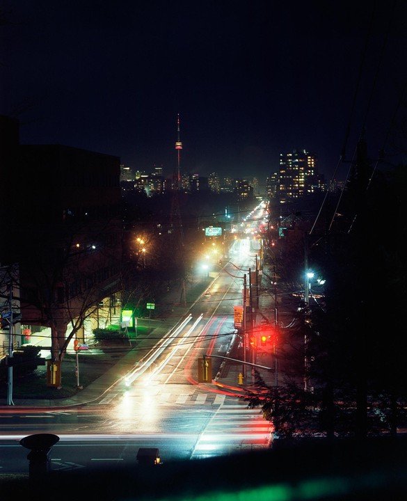 A long exposure shot captures light trails from vehicles on a city street at night, with the Toronto skyline and CN Tower visible in the distance.