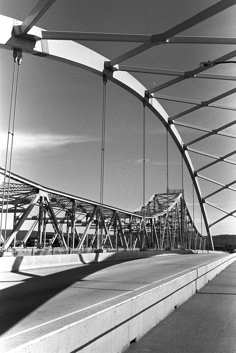 A black and white photograph captures the intricate geometric structure of a large bridge, with its arches and trusses creating strong shadows on the road surface.