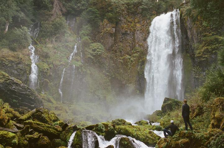 Two men stand and sit on mossy rocks looking at a large waterfall and smaller cascades in a lush, green forest.