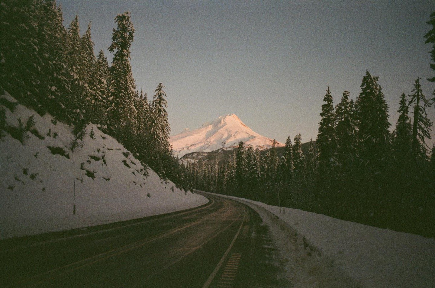 A winding road leads through a snowy forest towards a snow-capped mountain under a clear sky.