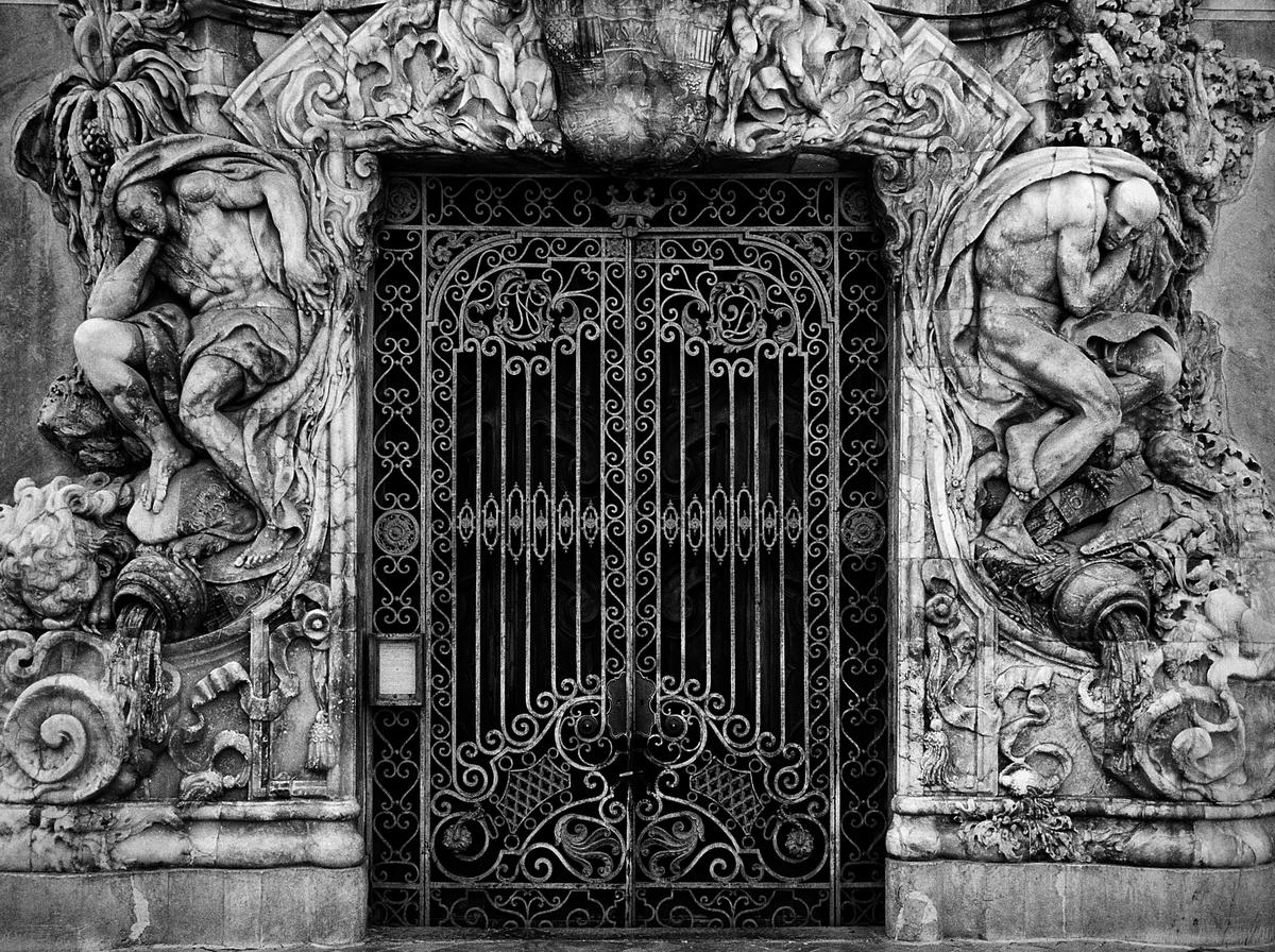 A black and white photo shows a highly ornate gate flanked by two large sculptures of men.