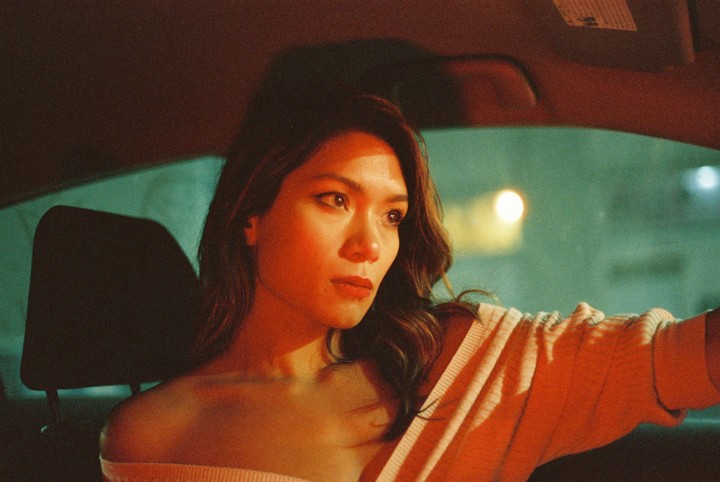 A woman with long hair sits in a car at night, illuminated by orange light.