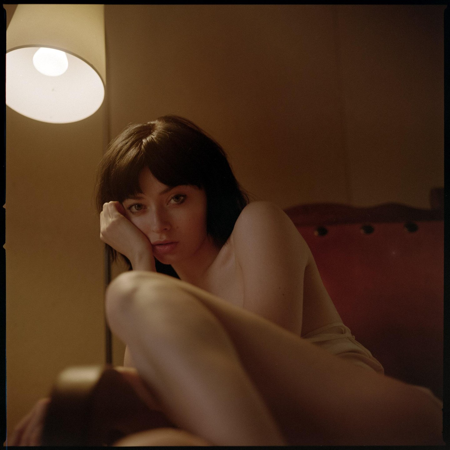 A woman with dark hair rests her head on her hand, looking directly at the viewer, with a lamp visible in the upper left.
