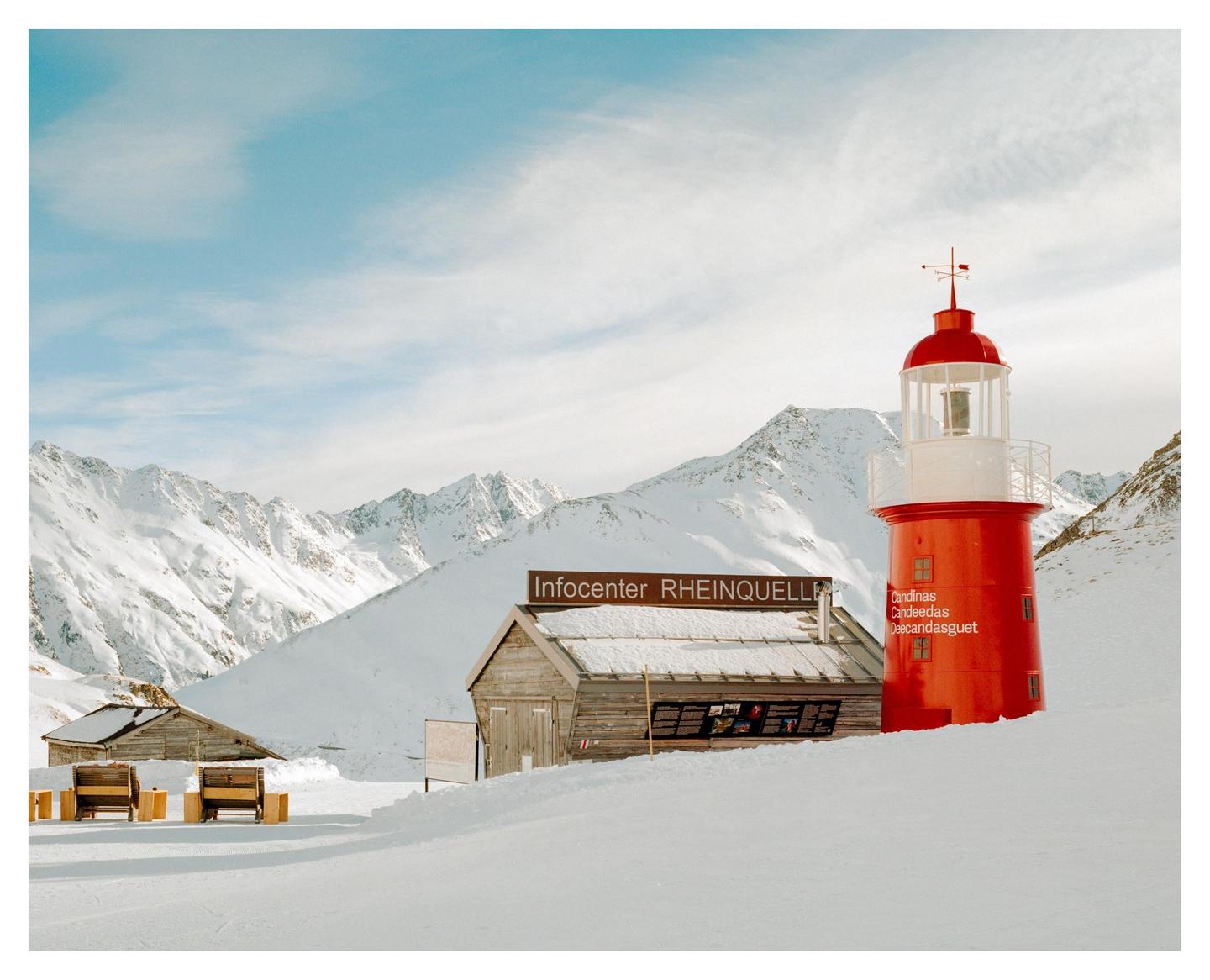 A red lighthouse stands next to a wooden infocenter building in a snowy mountain landscape.
