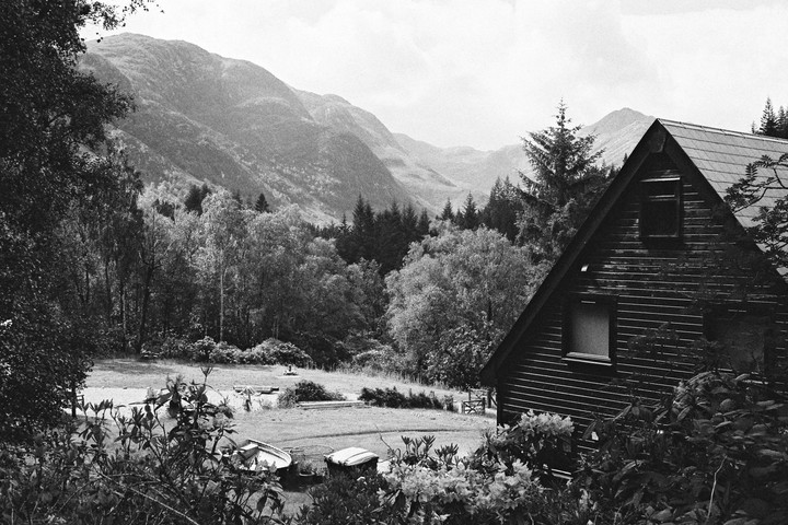A wooden cabin with a triangular roof is nestled among trees with mountains in the background.