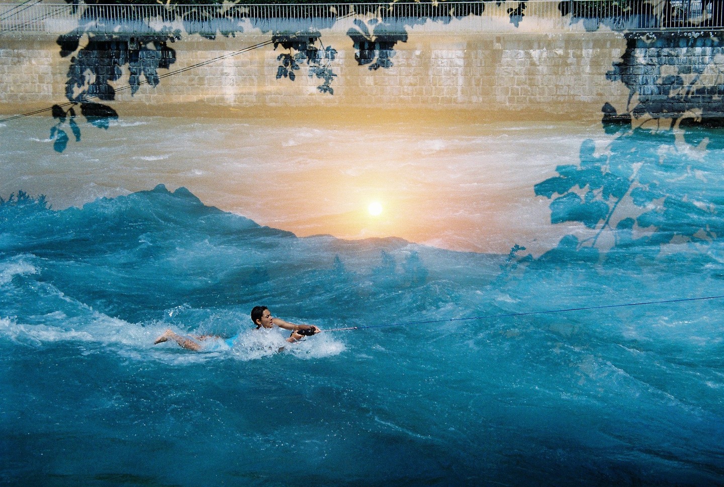 A woman is wakeboarding on a river with a sunset in the background and a wall with foliage in the foreground.