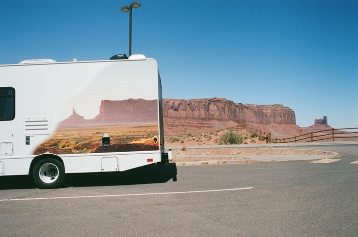 A white recreational vehicle with a desert landscape painted on its side is parked on asphalt with a desert landscape and blue sky in the background.
