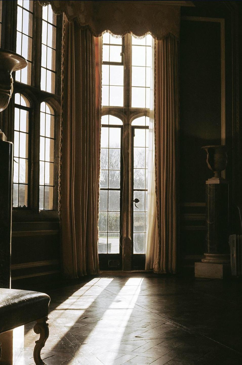 Sunlight streams through tall windows and a glass door into a dark room with a patterned floor.