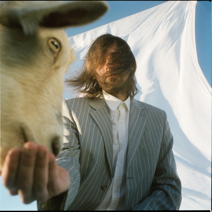 A man in a pinstripe suit stands next to a sheep, with a white sheet billowing in the background.