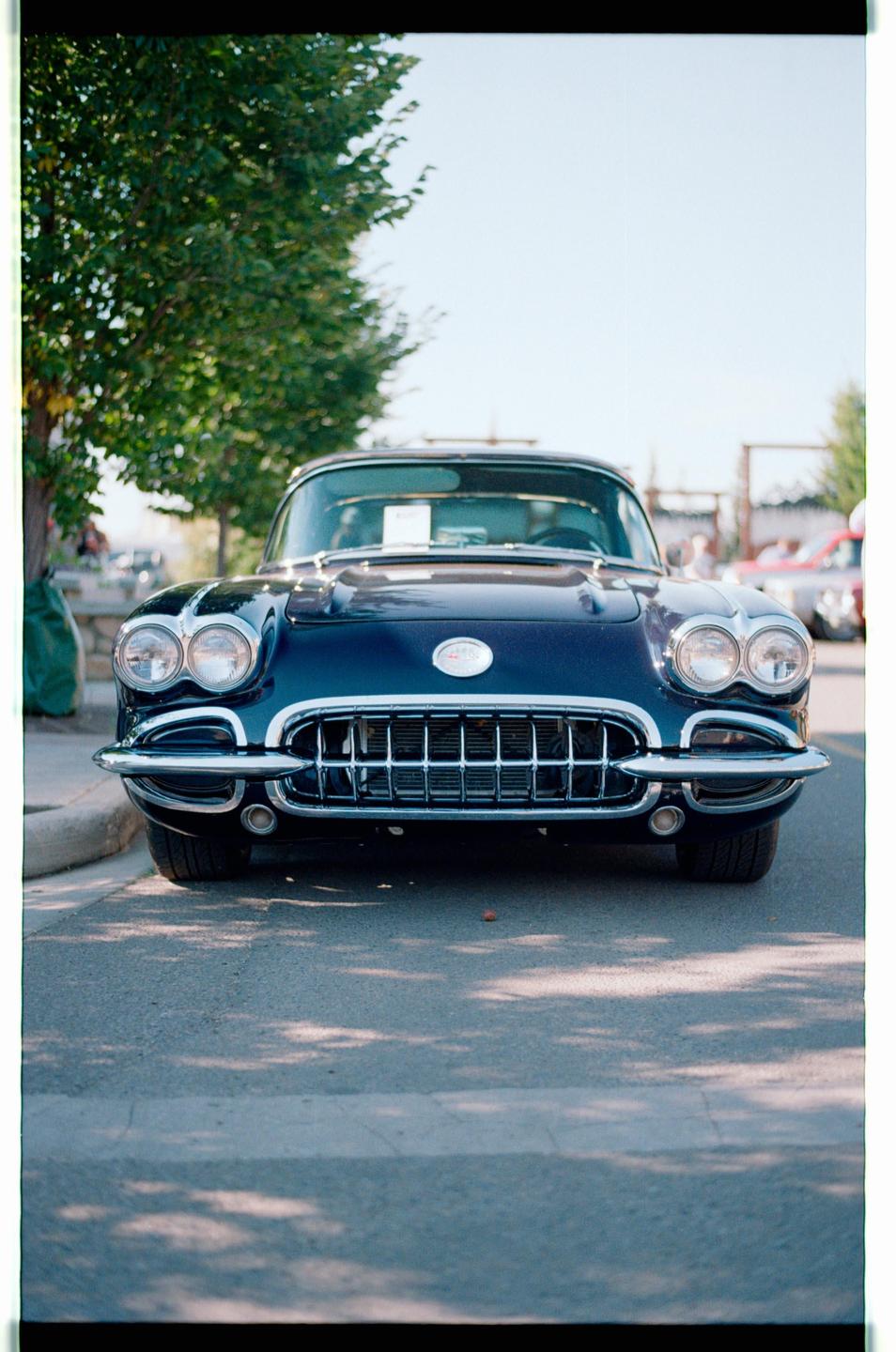 A dark blue vintage car with chrome details is parked on a street under dappled sunlight.
