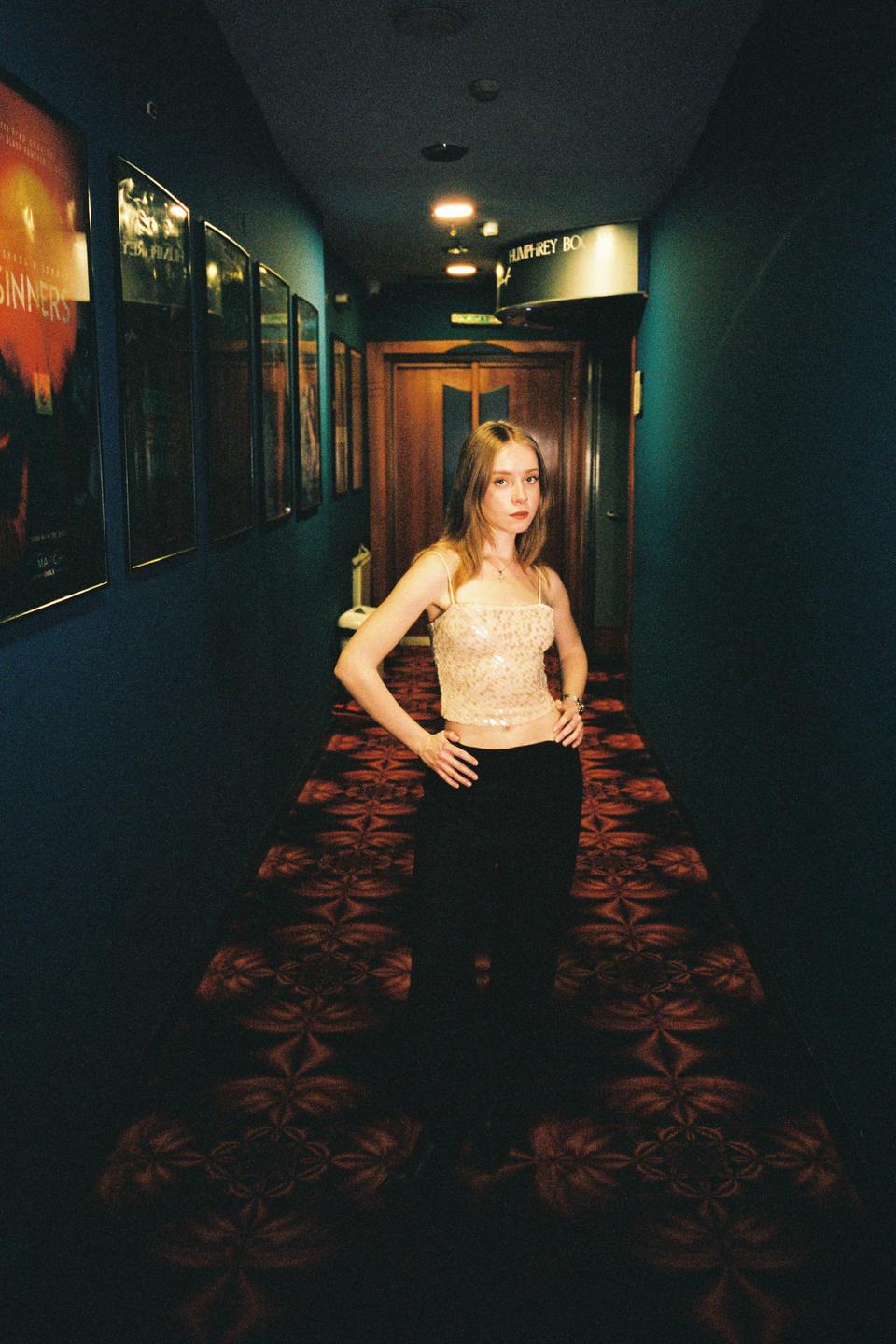 A woman stands in a dark hallway with movie posters on the wall and a patterned carpet.