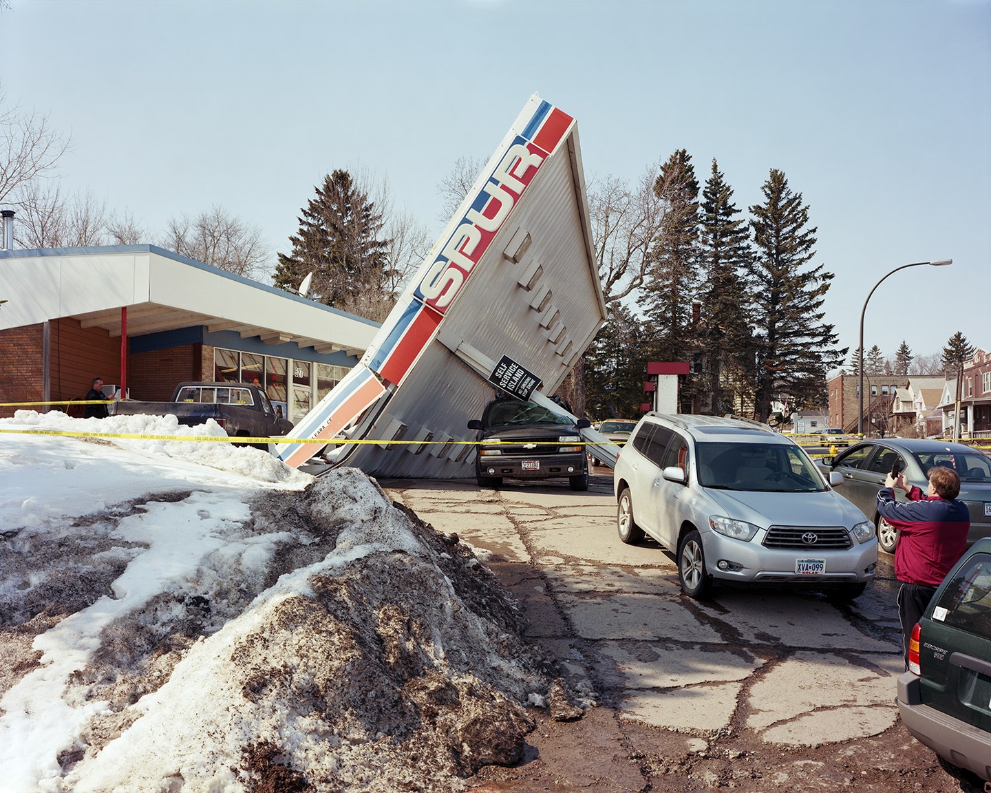 A gas station sign has fallen over onto a car, with yellow caution tape surrounding the area and a man taking a photo.