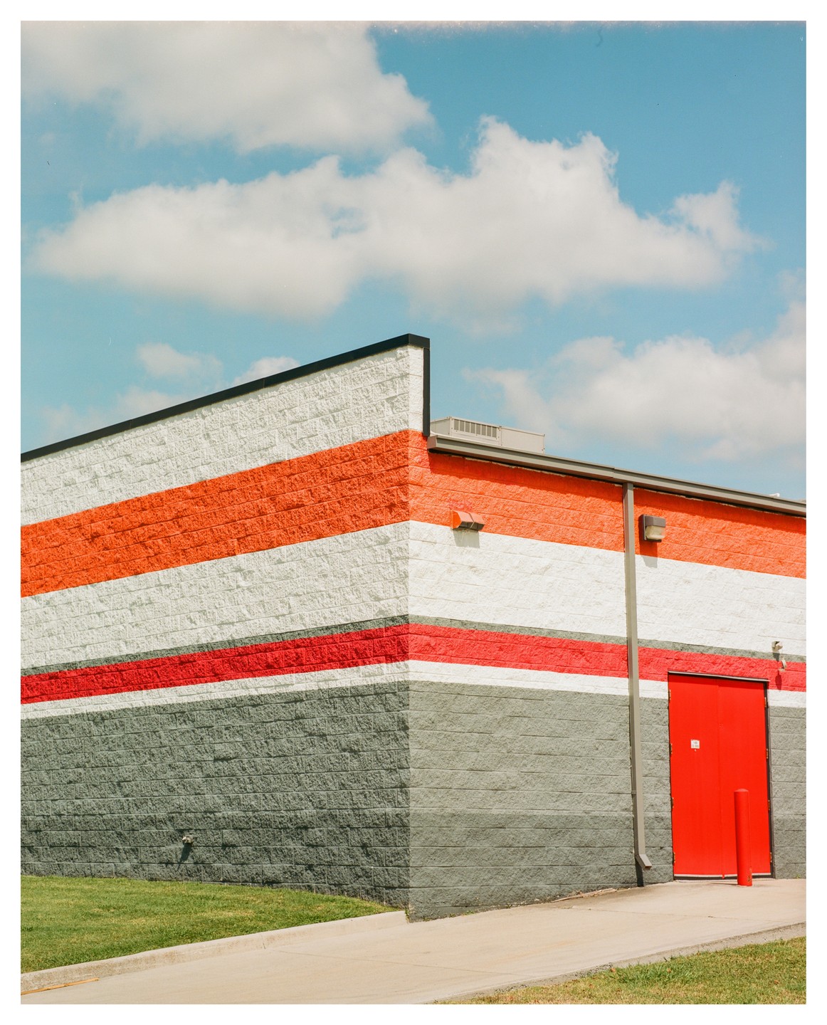 A building with orange, white, red, and gray stripes has a bright red door and a patch of green grass in front of it under a blue sky with clouds.