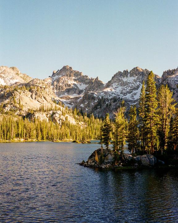 A lake is surrounded by a forest and snow-capped mountains under a clear sky.
