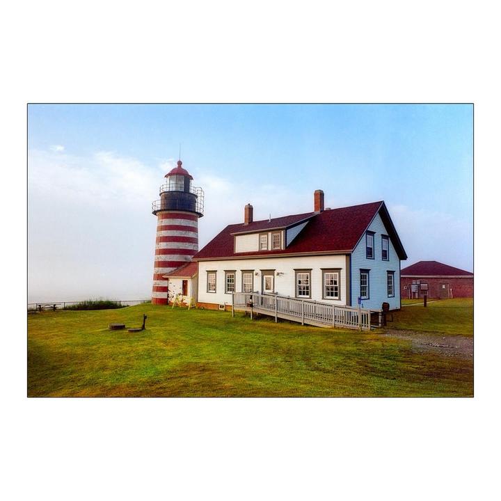 A red and white striped lighthouse stands next to a white house with a red roof on a grassy hill under a hazy sky.
