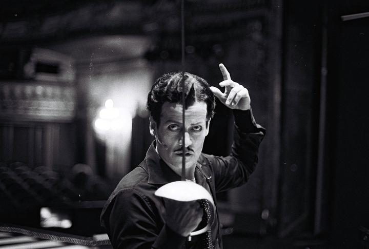 A man with a painted face and a mustache holds a fencing foil and points his finger up.