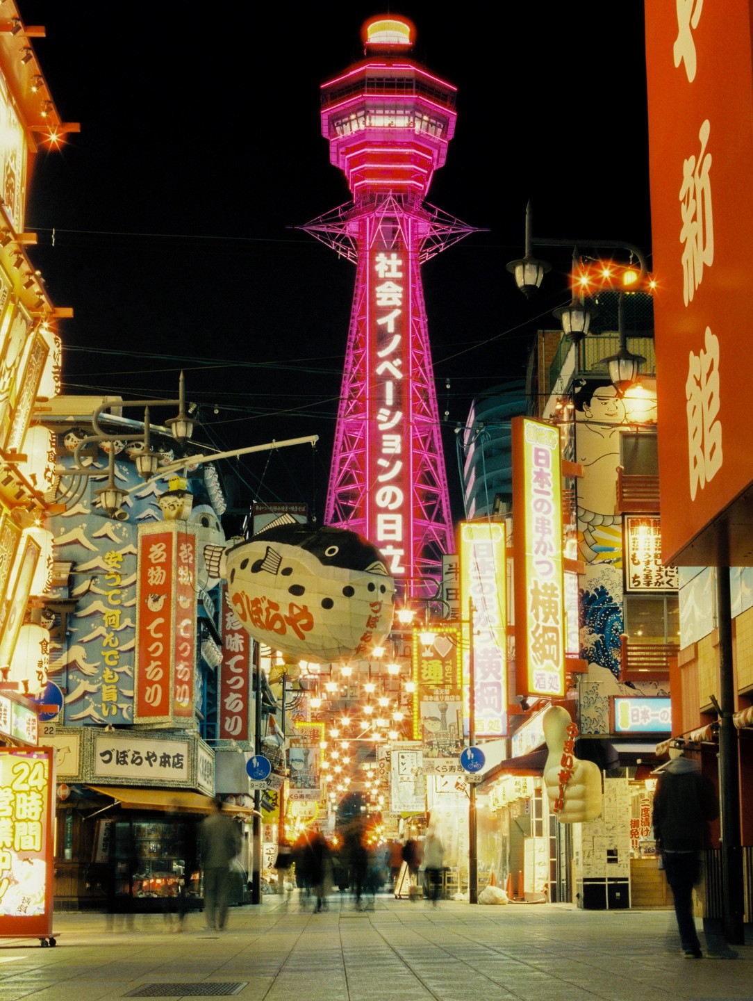 A street at night is illuminated by neon signs and a tall, pink tower.