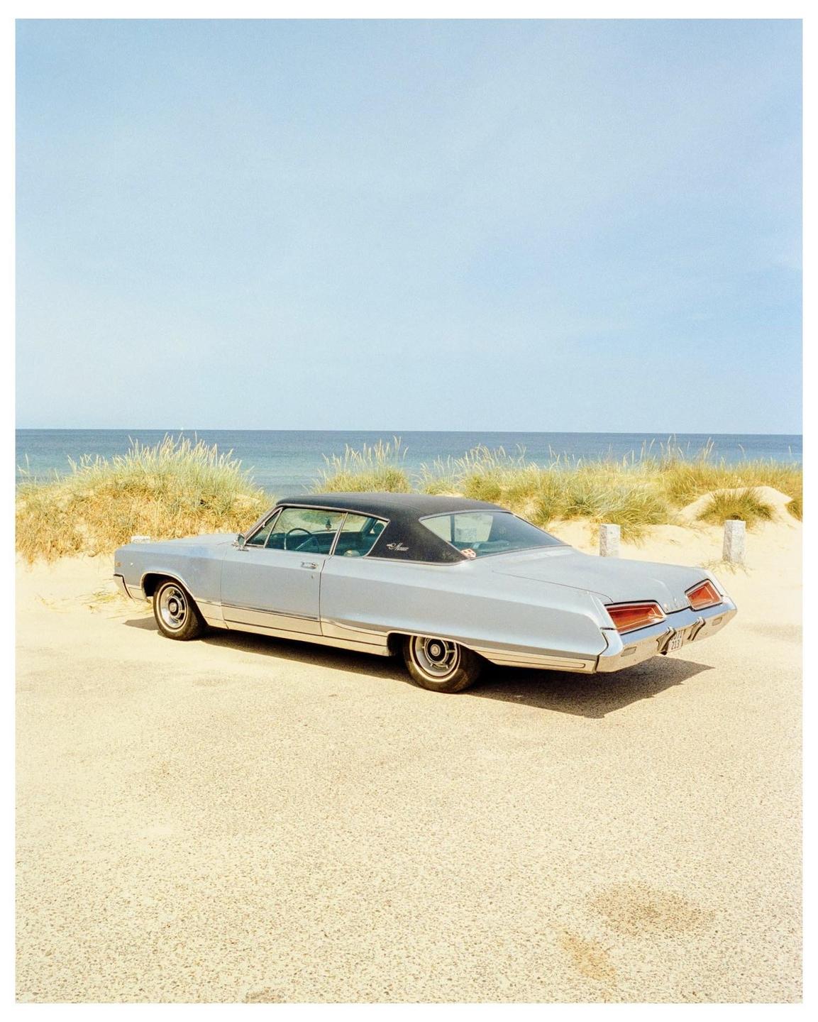 A light blue vintage car with a black roof is parked on a sandy road next to a beach with tall grass and the ocean.