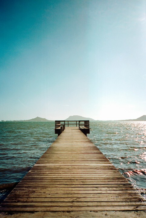 A wooden pier extends into a body of water with mountains in the distance under a clear sky.
