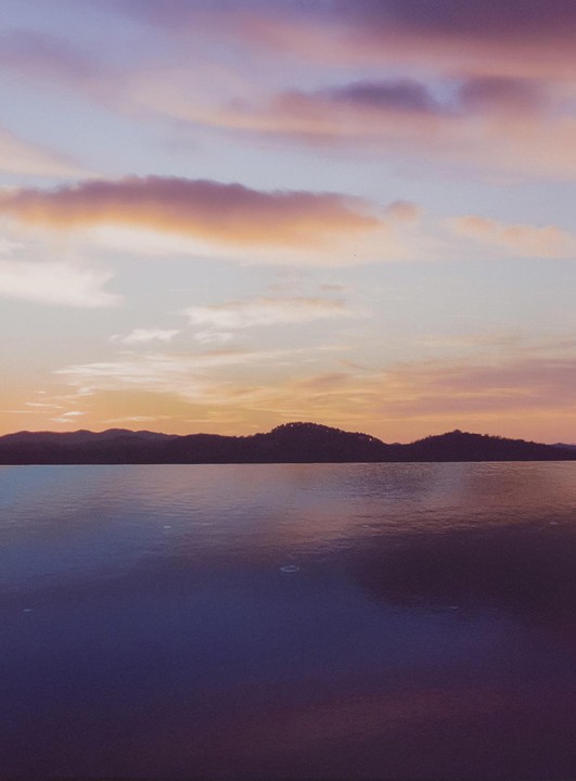 A body of water reflects the colorful sky and silhouetted hills at sunset.
