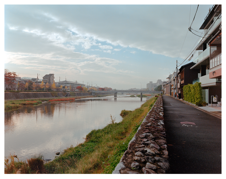 A river flows past a stone wall and a street lined with buildings and trees with autumn foliage.