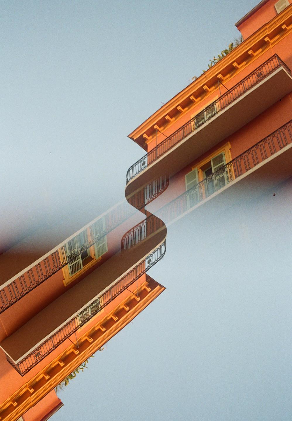 A double exposure shows an orange building with balconies against a light blue sky, creating a wavy, mirrored effect.