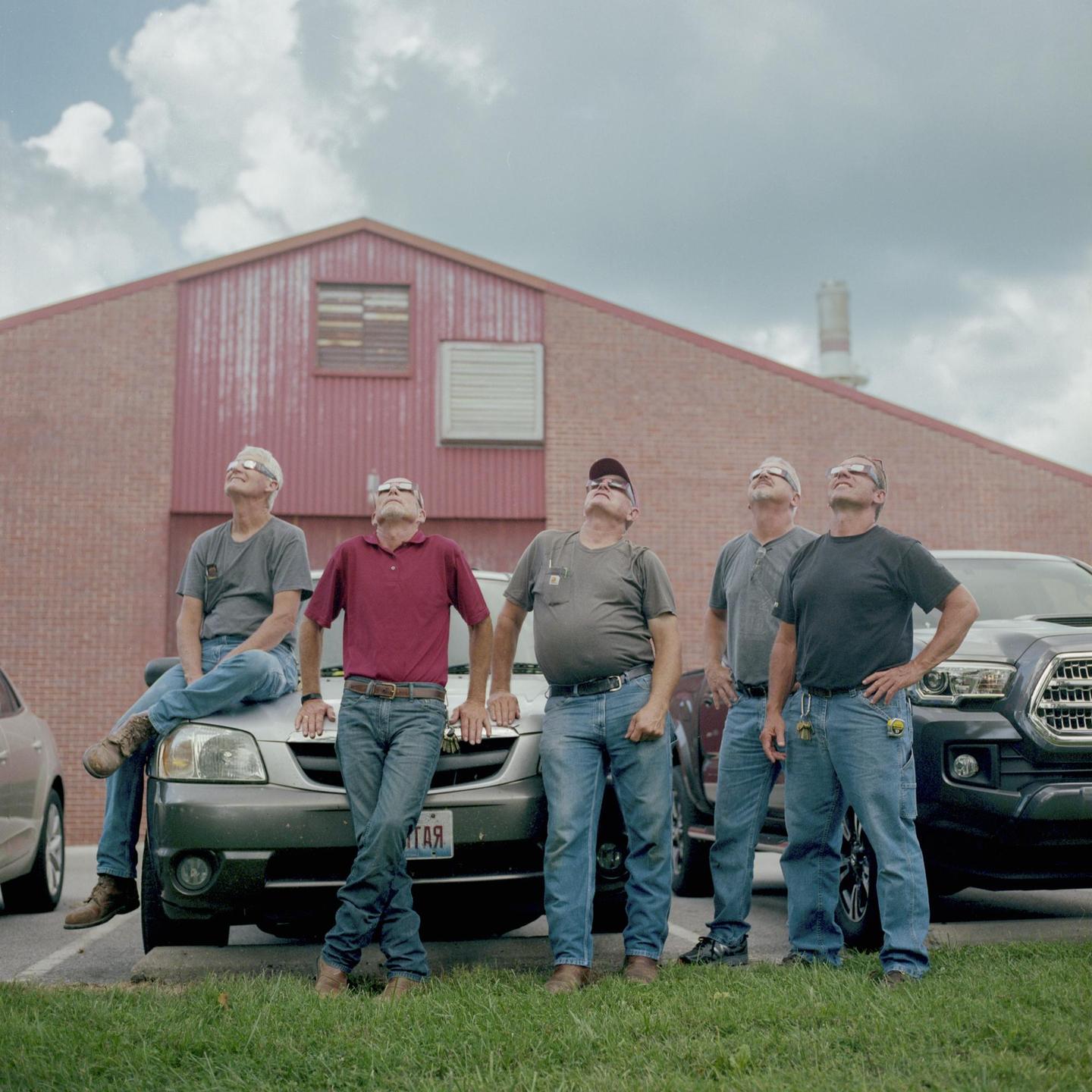 Five men wearing protective glasses look up at the sky in front of a red brick building.