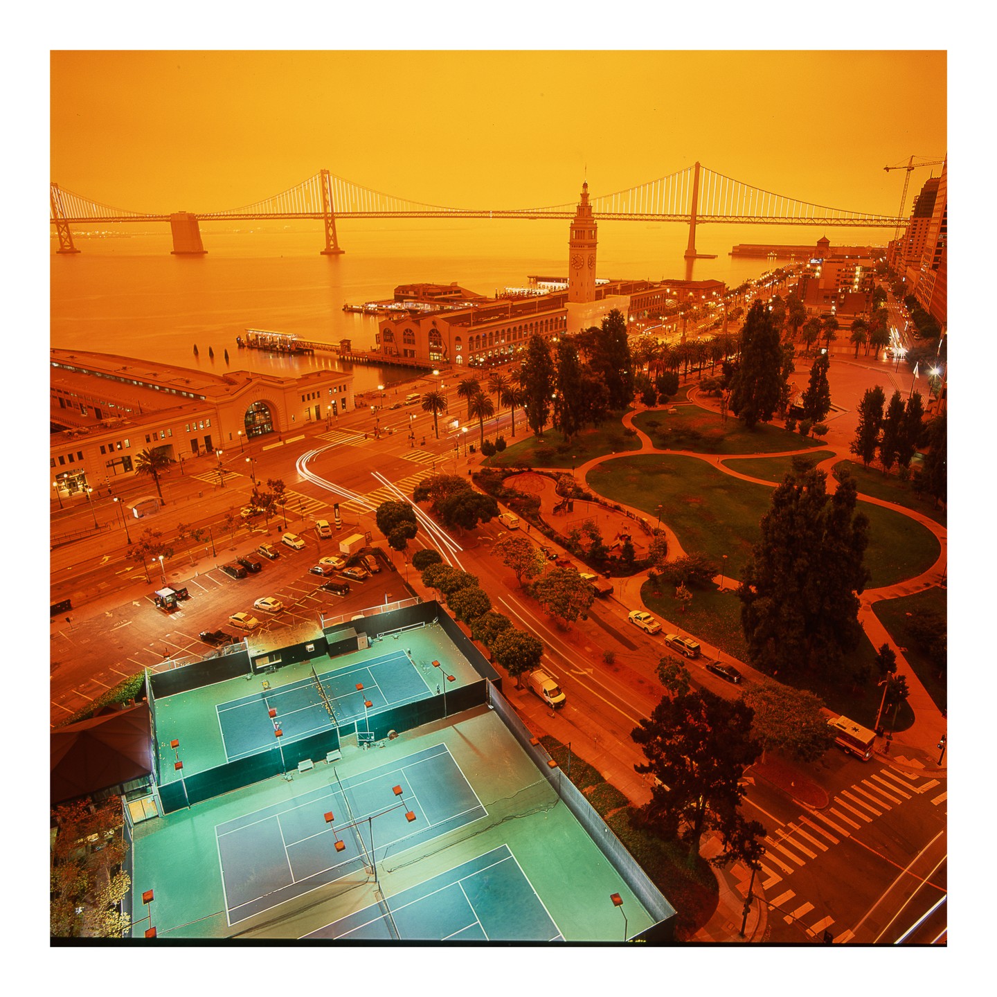 An aerial view shows tennis courts, a park, a street with cars, and a bridge over water under an orange sky.