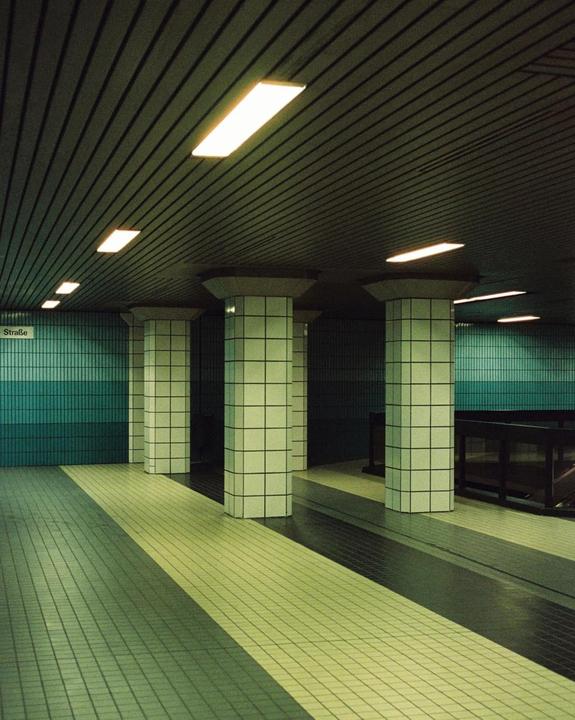A subway station platform features tiled walls and columns, with fluorescent lights illuminating the ceiling.