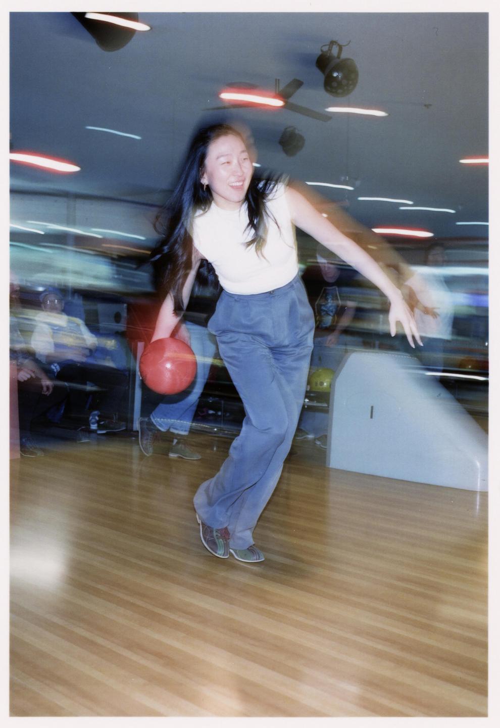 A woman smiles while bowling a red ball down a lane in a blurry bowling alley.