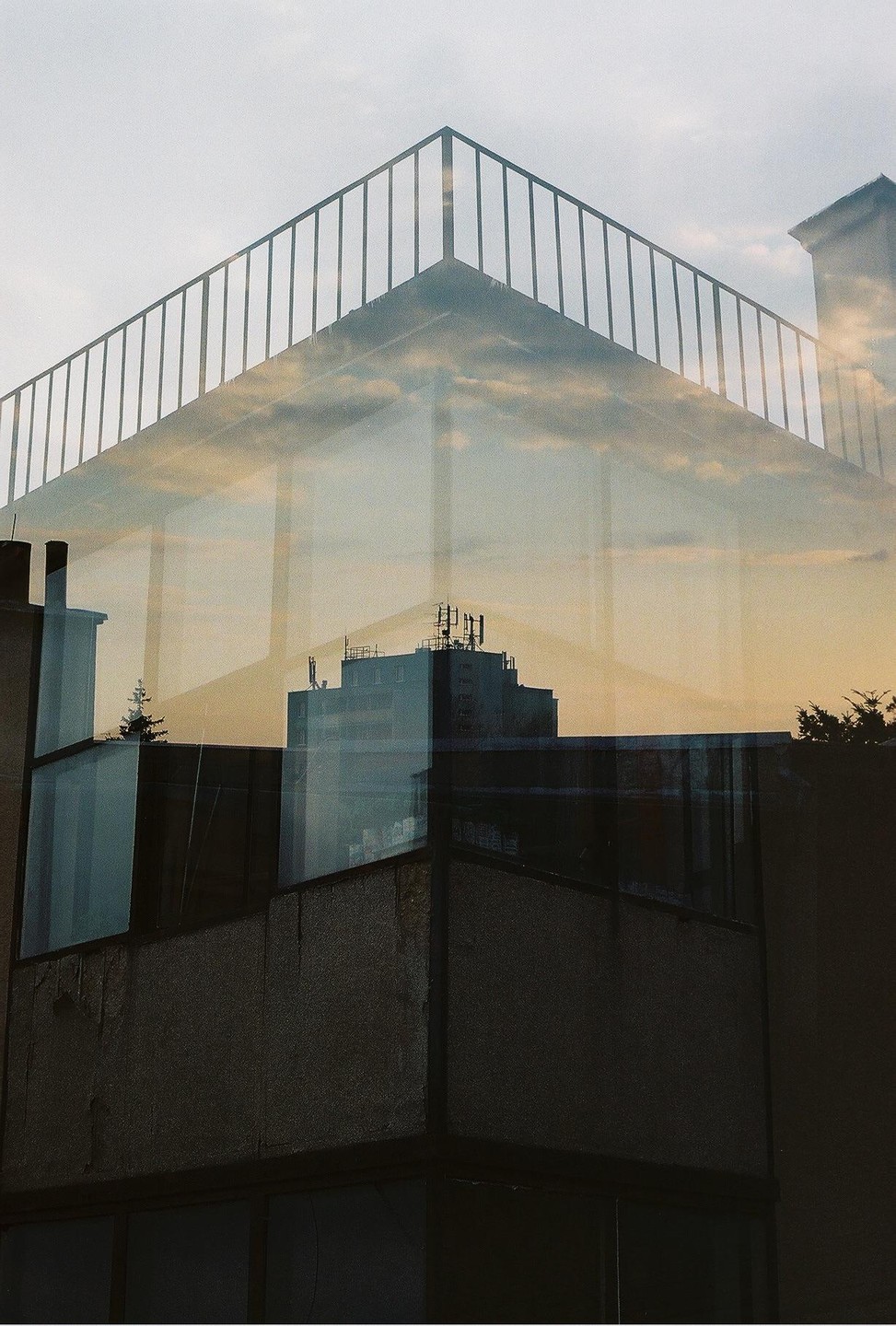 A double exposure shows a building with a railing on top and a reflection of clouds and another building.
