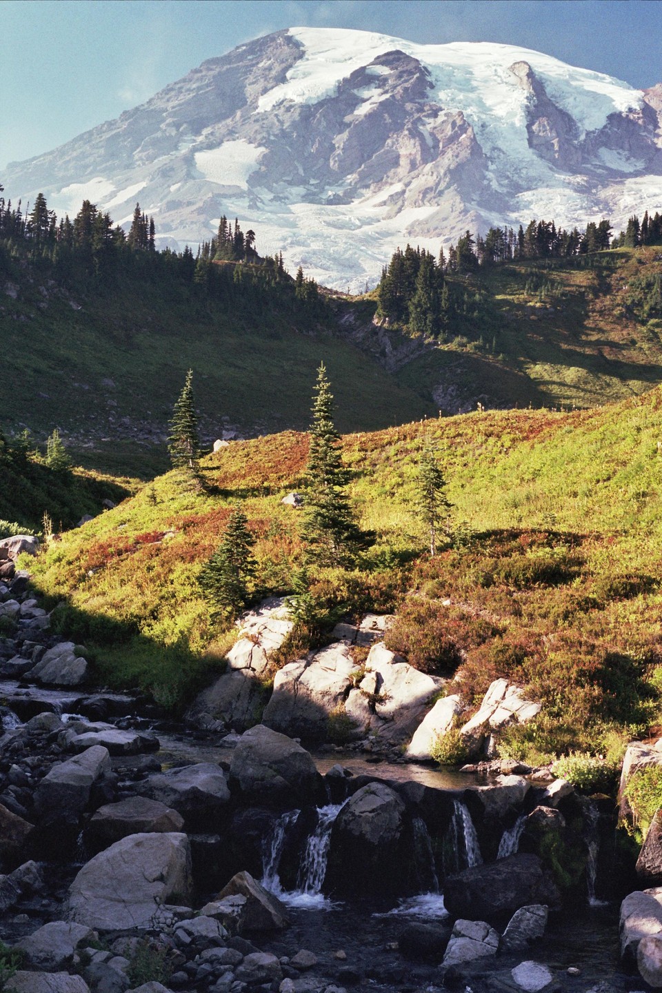 A snow-capped mountain rises above a valley with a rocky stream and evergreen trees.