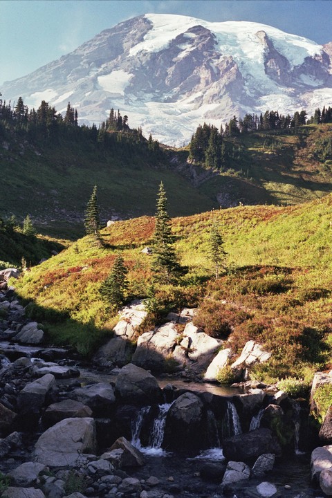 A snow-capped mountain rises above a valley with a rocky stream and evergreen trees.