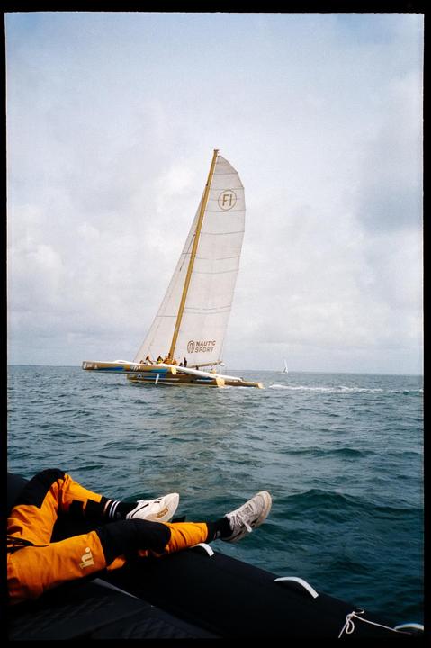A person's legs are visible in the foreground of a body of water with a sailboat in the distance.