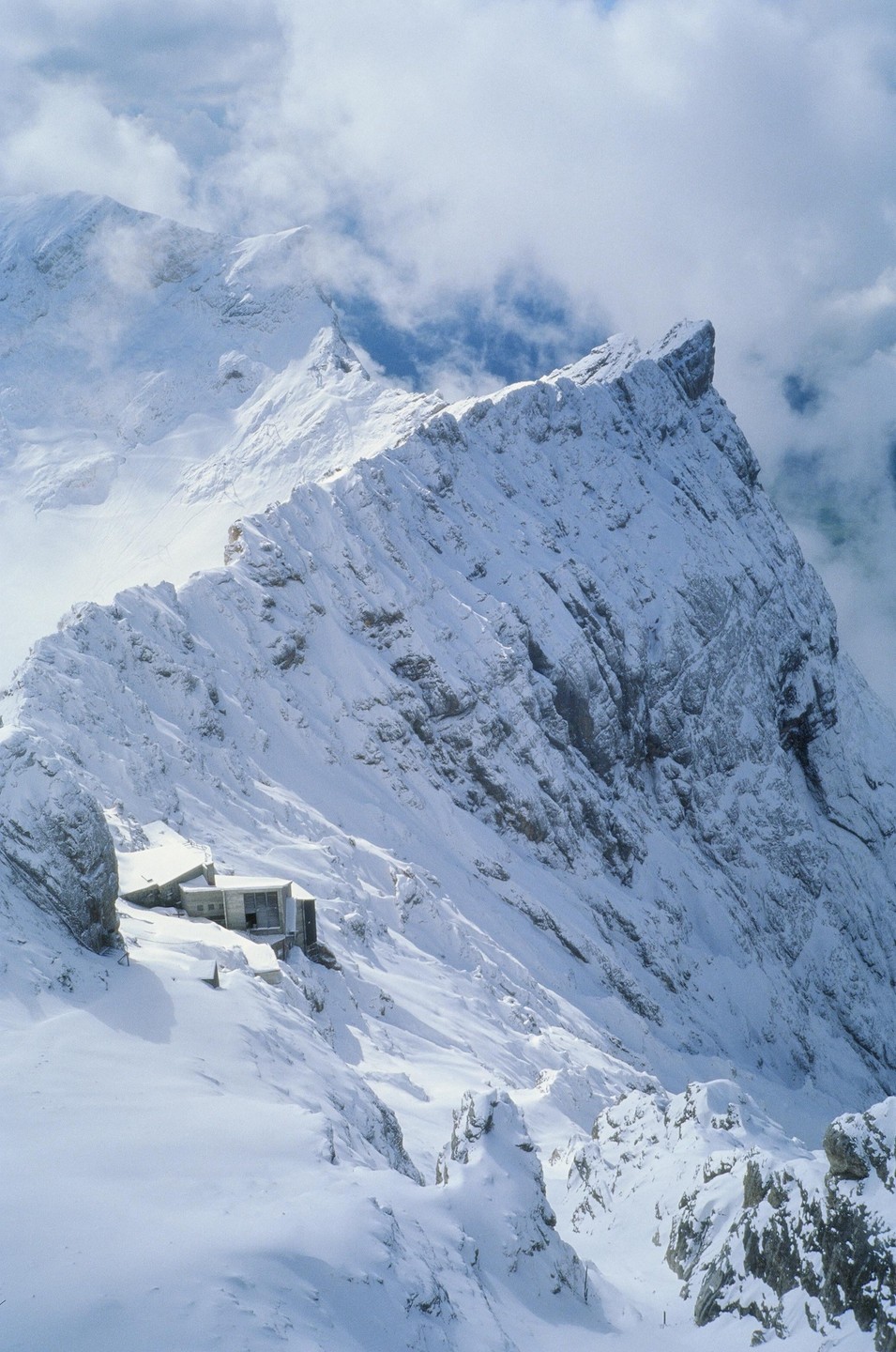 A building is nestled into the side of a snow-covered mountain with clouds in the background.