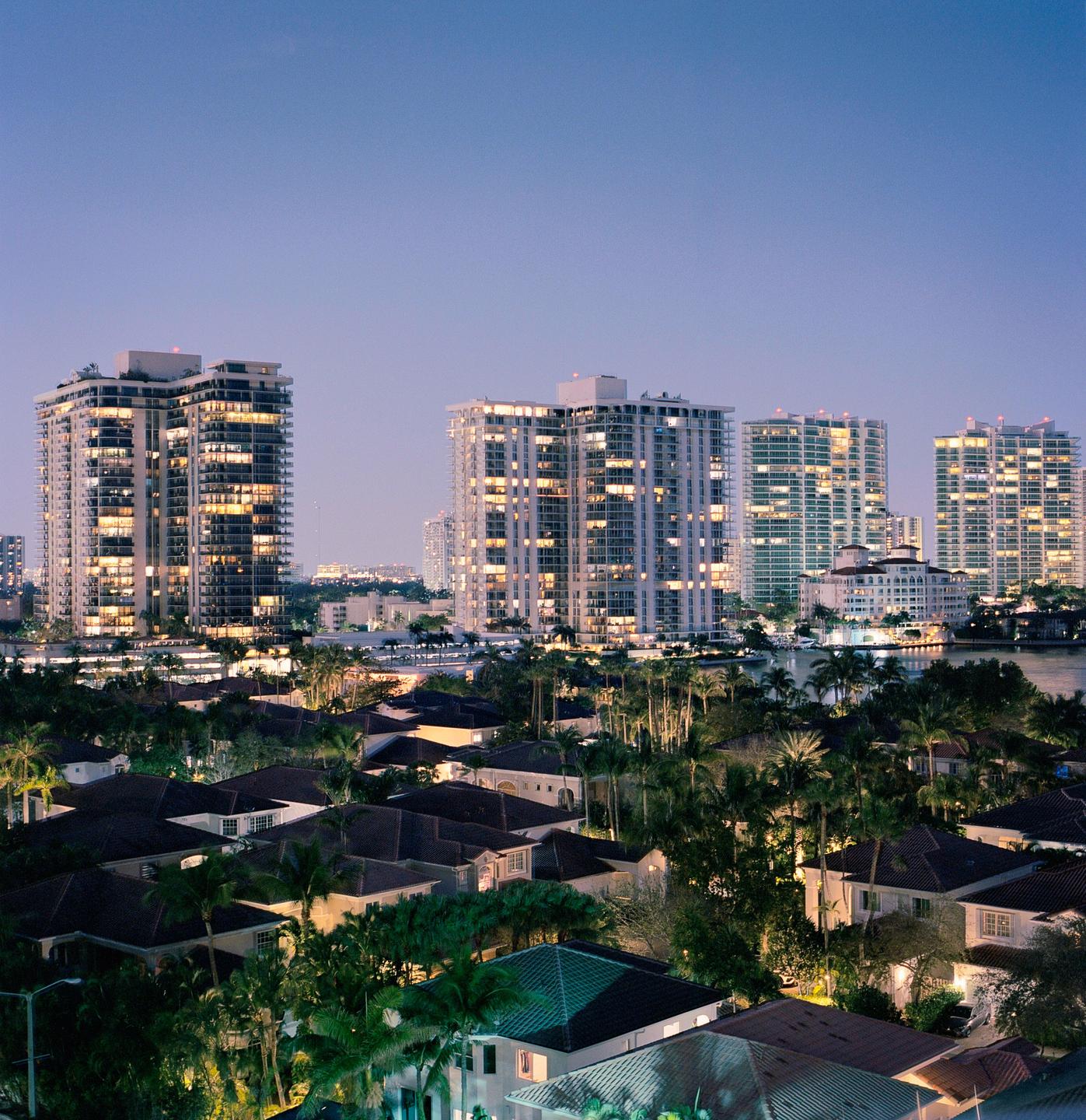 A city at dusk shows residential buildings and high-rises with illuminated windows.