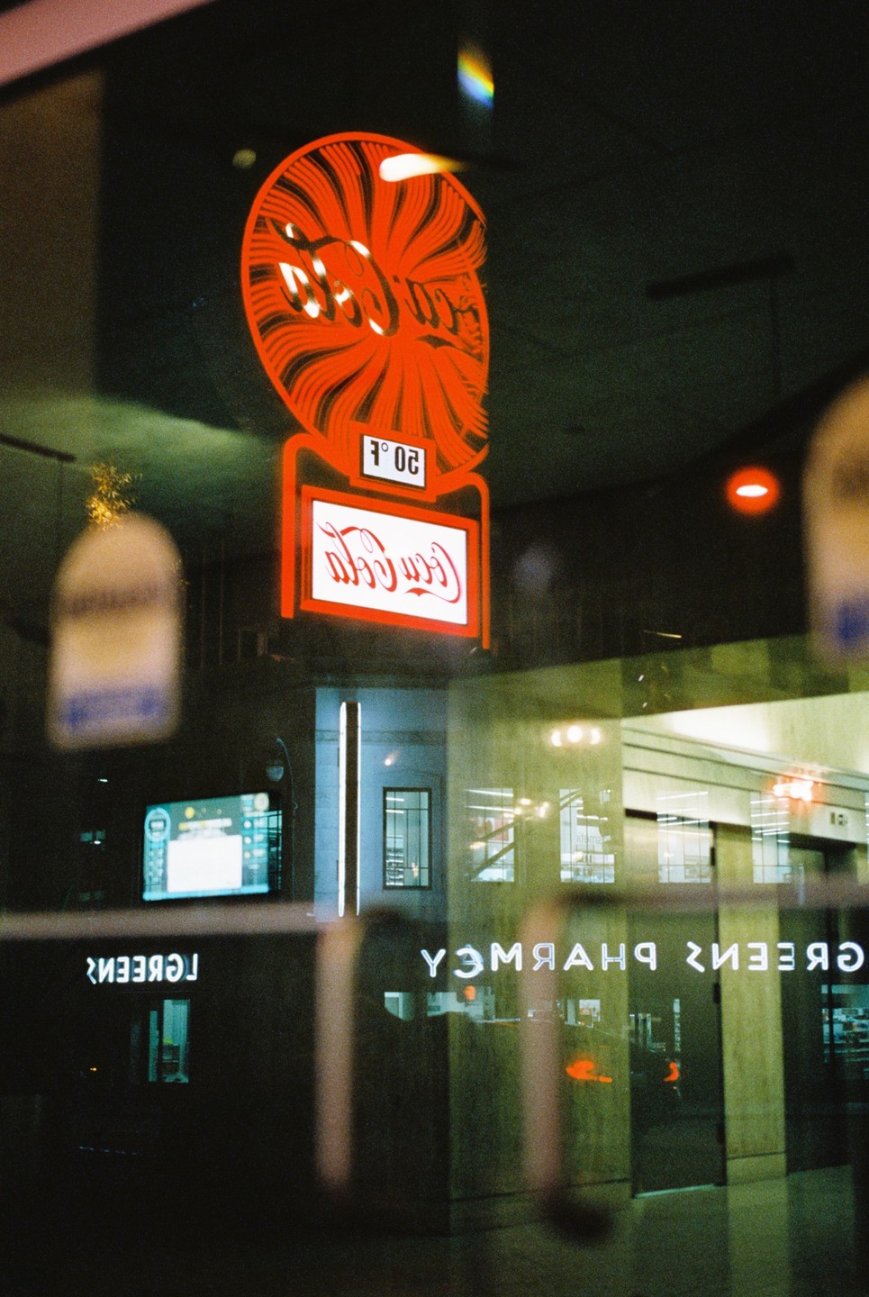 A red Coca-Cola sign is reflected in a window at night, with other neon signs and building interiors also visible in the reflection.