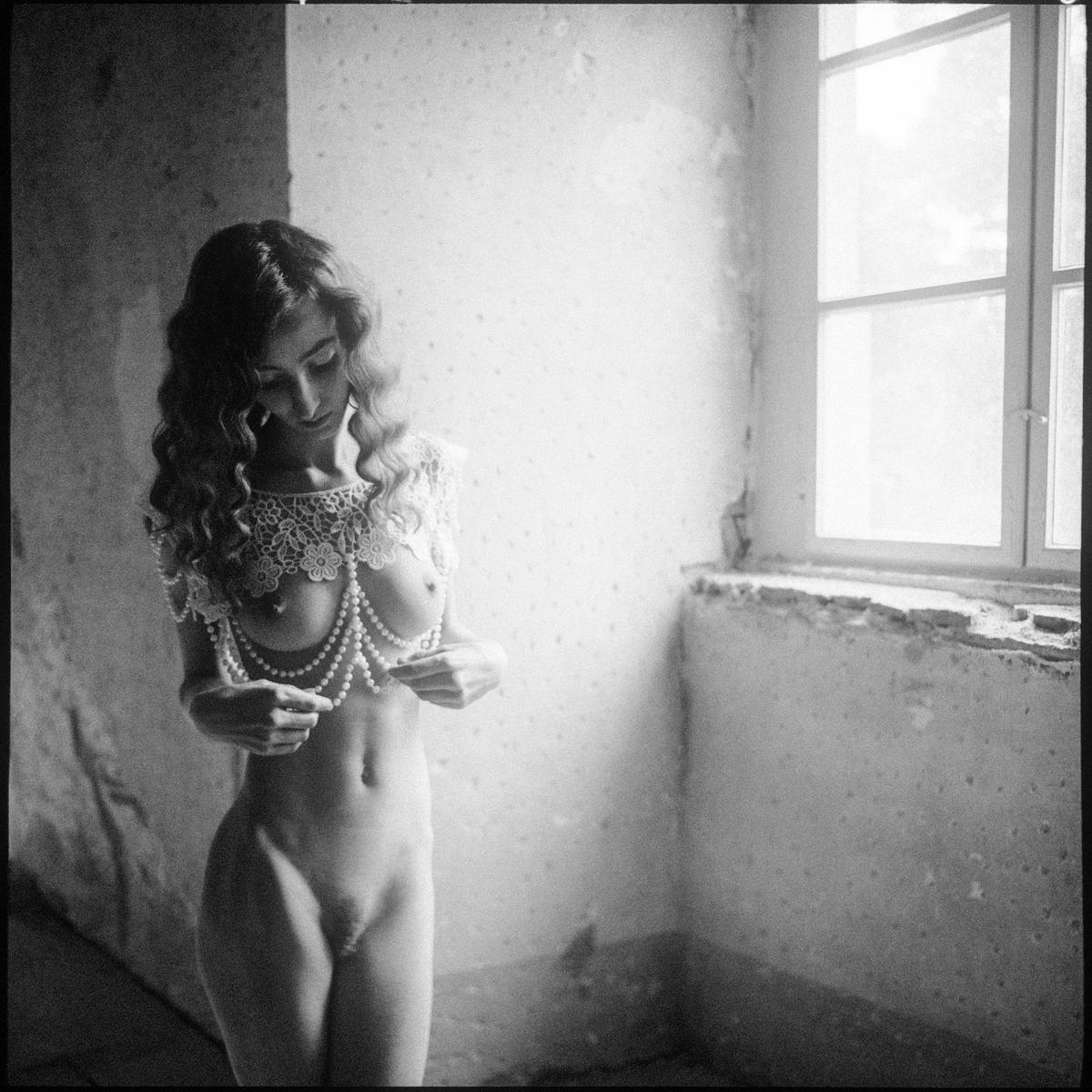 A nude woman with long curly hair stands in a room, adorned with a lace and pearl necklace, looking down at the pearls in her hands.