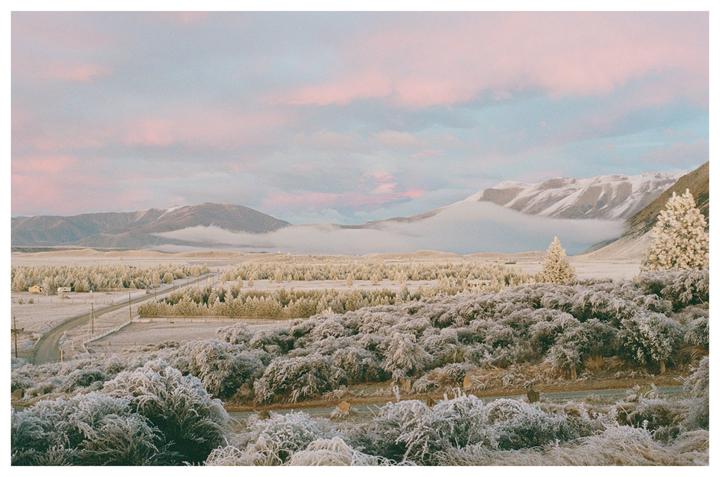 A winter landscape shows a road winding through a valley with snow-covered trees and mountains under a pink and blue sky.