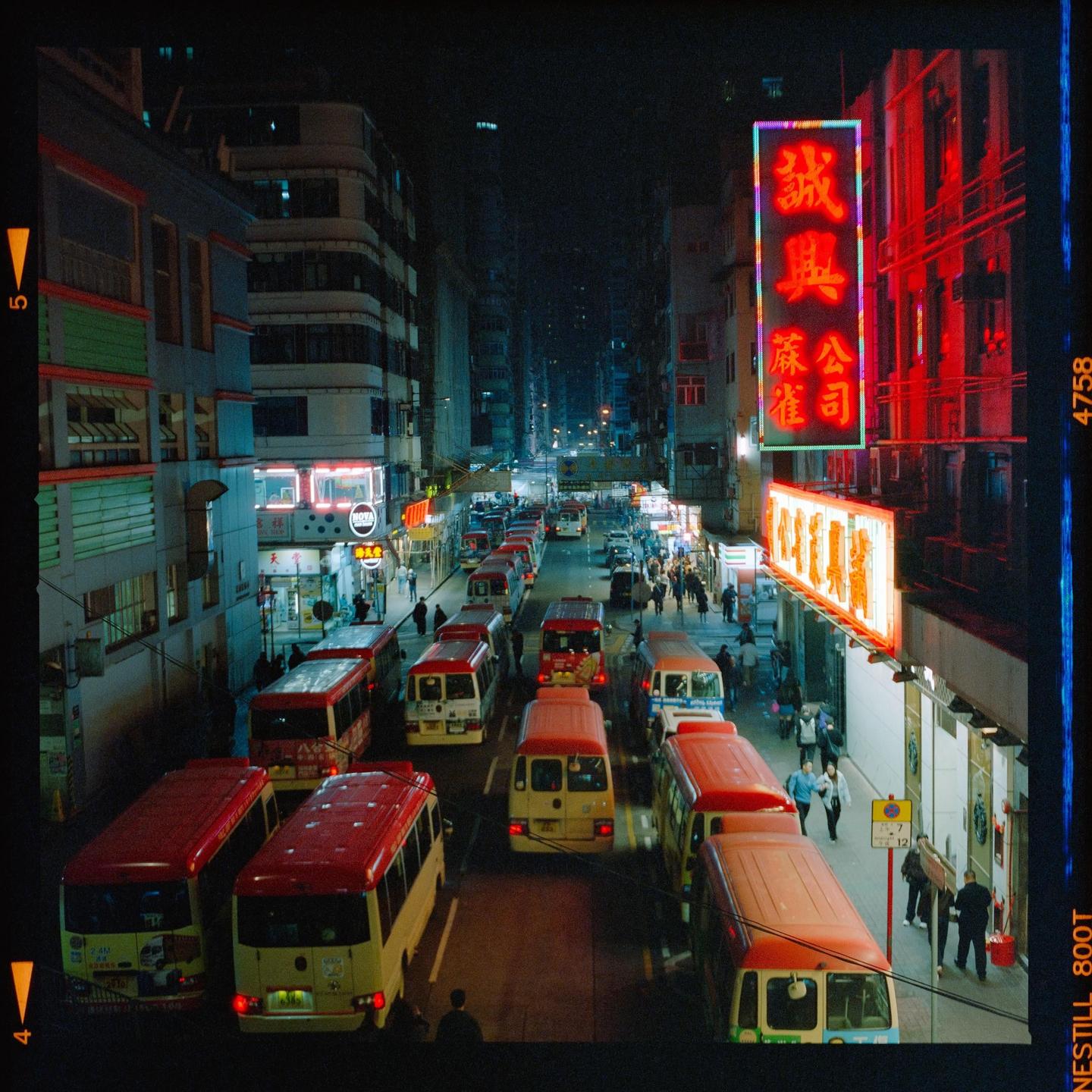 A street in Hong Kong is filled with traffic and pedestrians at night, with neon signs illuminating the buildings.