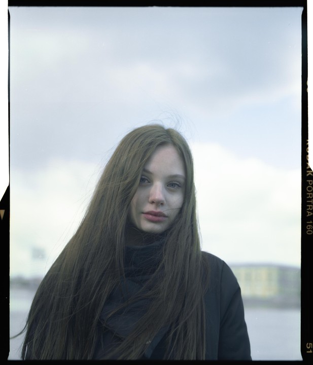 A woman with long hair and a black turtleneck looks directly at the viewer with a cloudy sky in the background.
