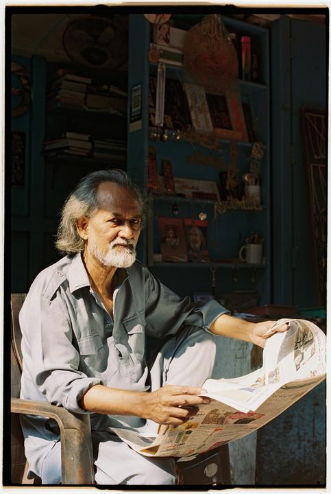 A man with a beard and long hair sits in a chair, holding a newspaper.