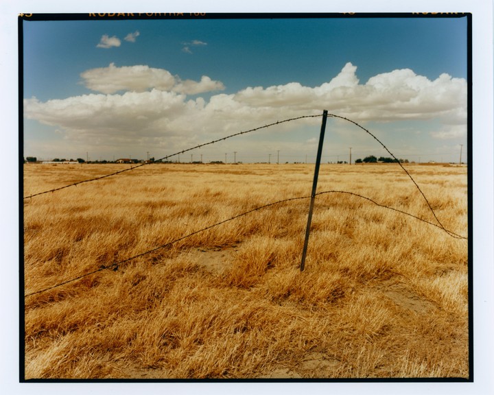 A barbed wire fence runs through a field of dry grass under a blue sky with white clouds.