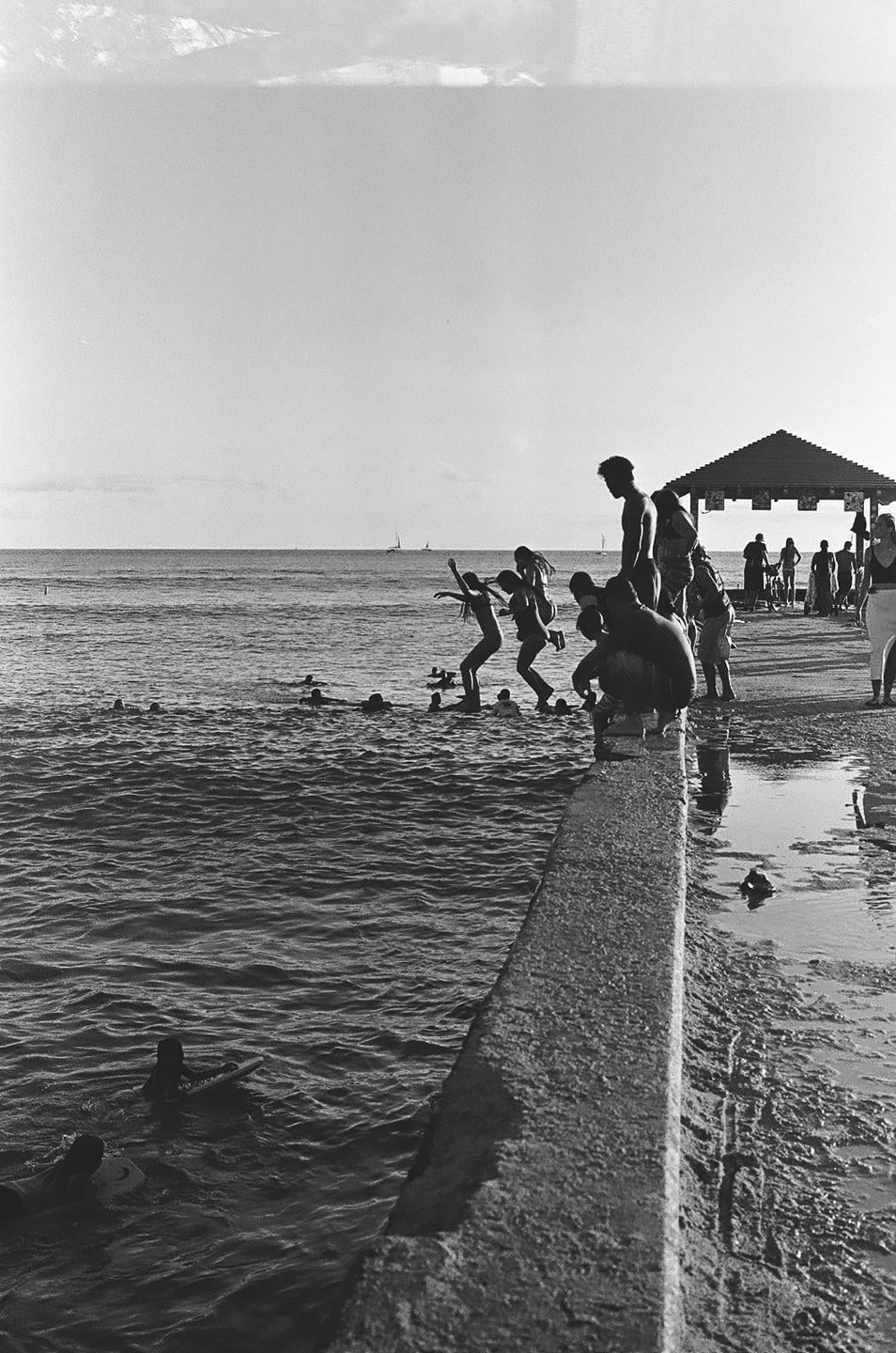 Several people are jumping into the ocean from a concrete pier, while others stand on the pier or swim in the water.