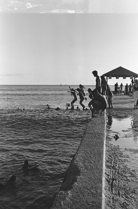 Several people are jumping into the ocean from a concrete pier, while others stand on the pier or swim in the water.