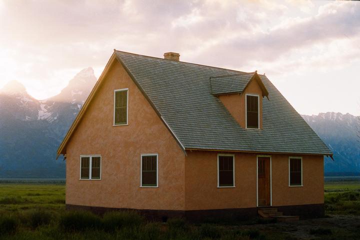 A house with a gray roof and orange walls stands in a field with mountains in the background under a cloudy sky.