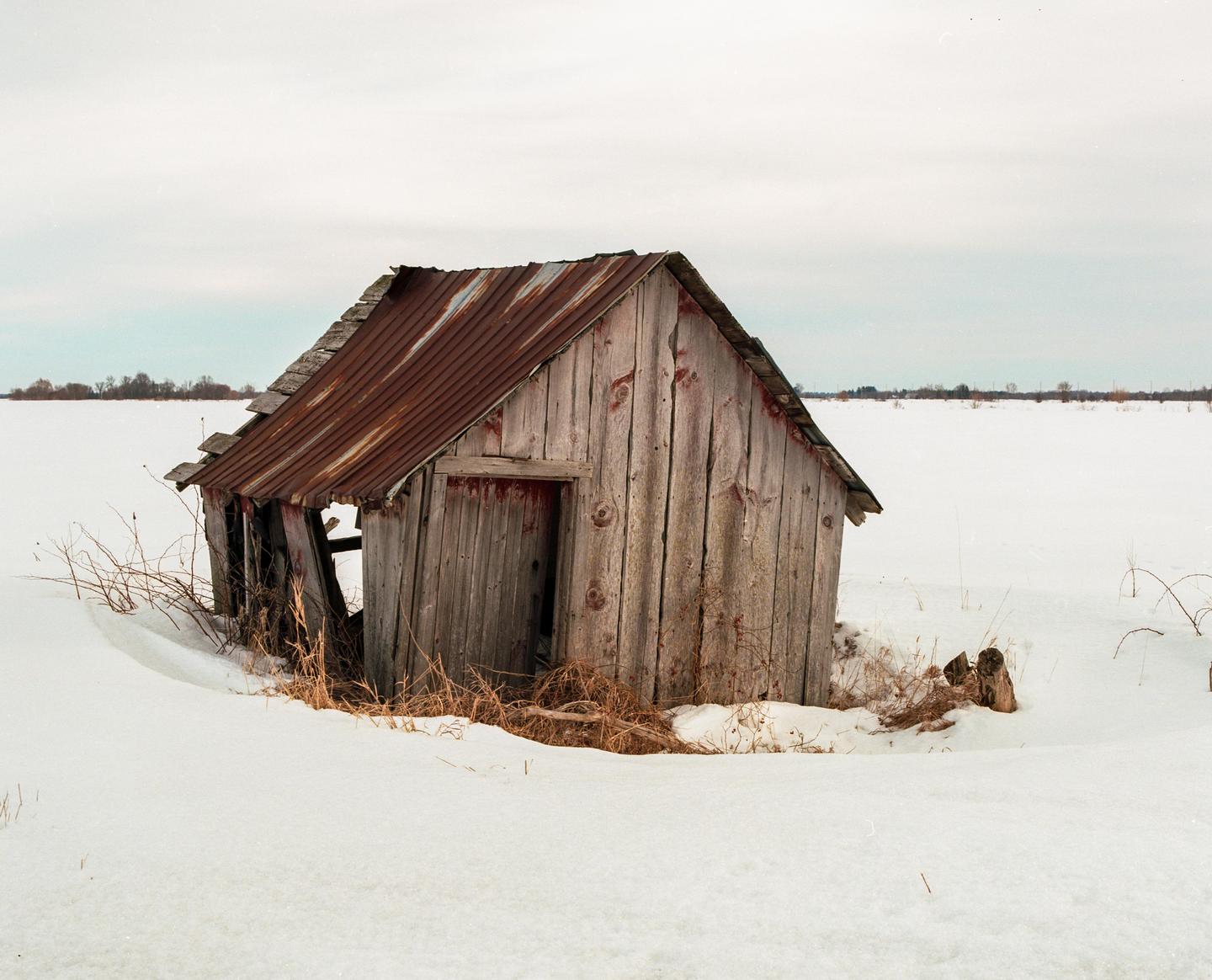 A weathered wooden shack with a rusty metal roof stands in a snow-covered field under a cloudy sky.